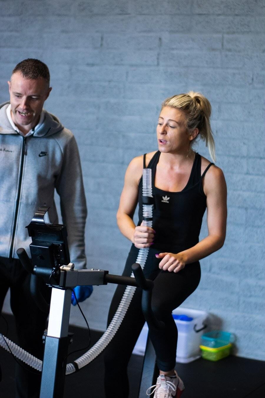 Fitness professional alongside woman sitting on a stationary bicycle with a VO2 tube in her hand as she exhales