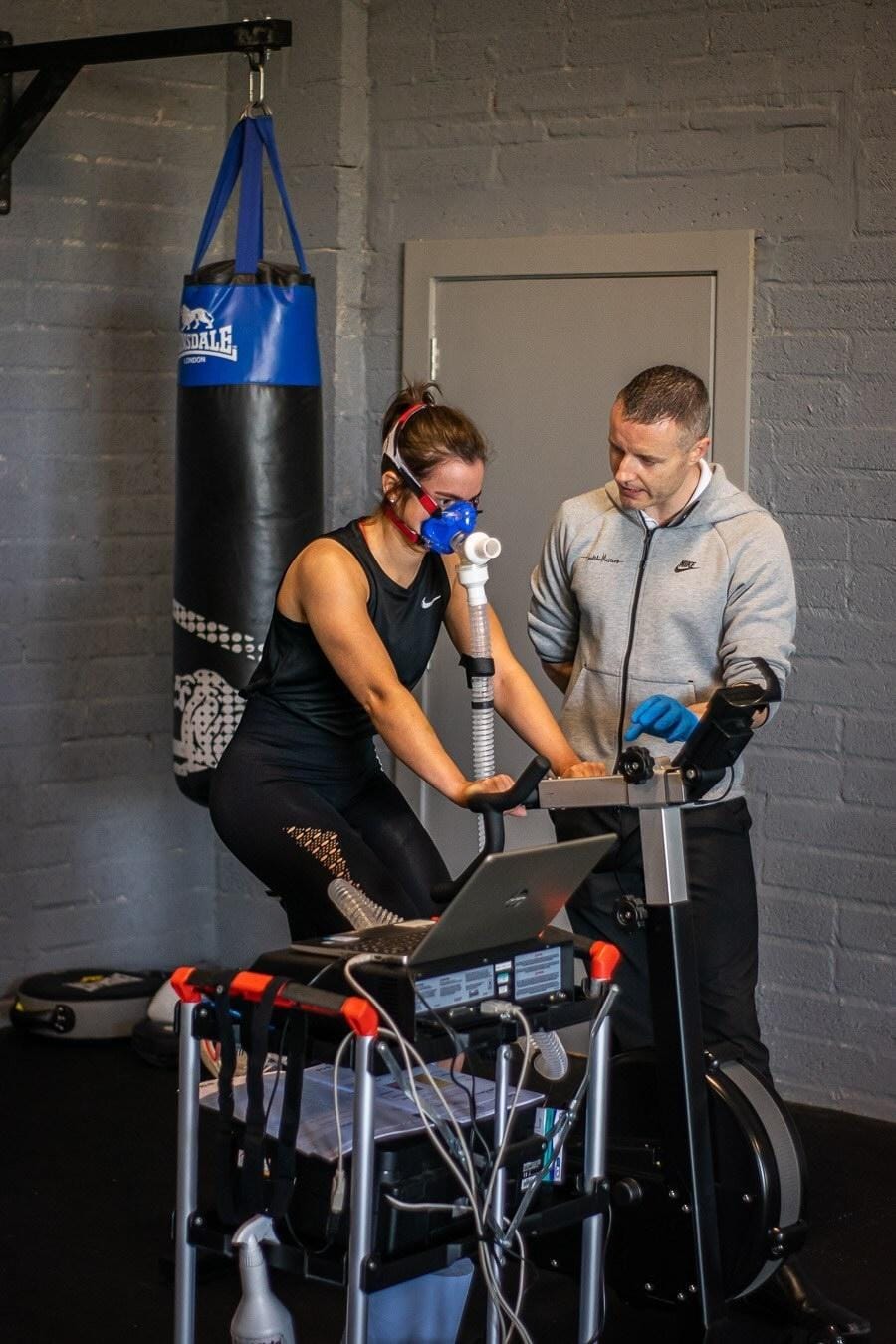 Fitness professional alongside woman sitting on a stationary bicycle with a VO2 tube and mask attached to her face.